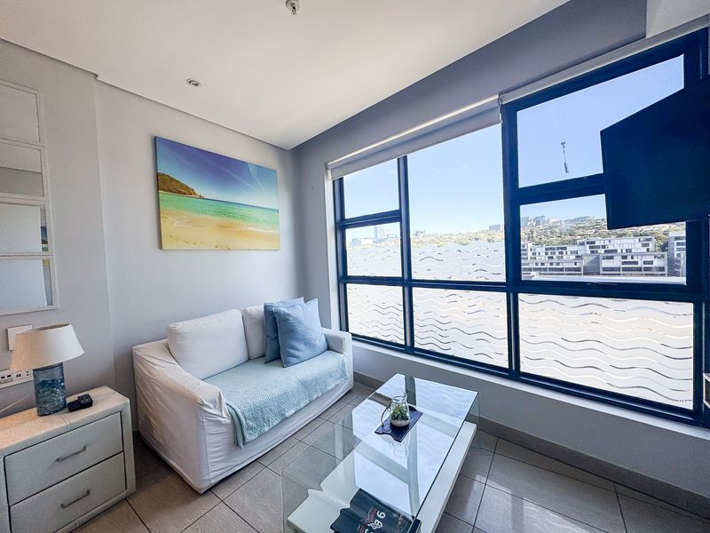 A bright living room featuring large aluminium-framed windows with city views, tiled flooring, and modern recessed lighting.