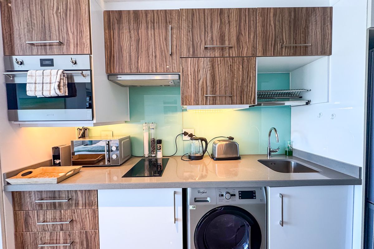 Modern compact kitchen featuring wood-grain and white cabinetry, stone countertops, glass splashback, built-in oven, and an integrated washing machine.