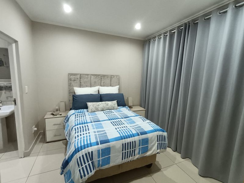 A well-maintained bedroom featuring neutral tiled flooring, recessed downlighting, and full-length curtains, with an adjoining bathroom visible.