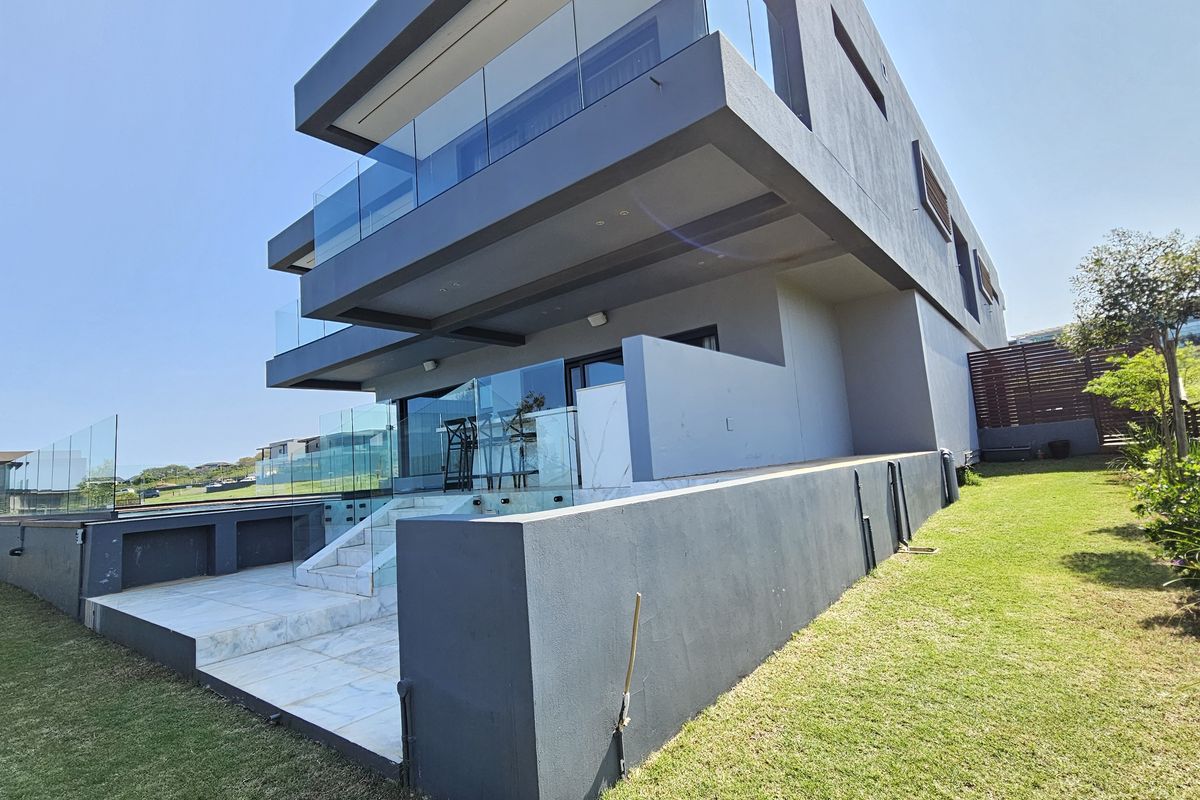 Modern multi-story rear elevation featuring glass balustrades, a tiled patio area, and clean architectural lines overlooking a landscaped garden.