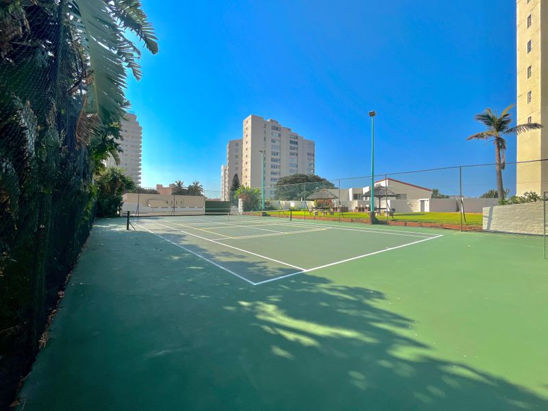 A well-maintained outdoor tennis court facility featuring perimeter fencing and floodlights, situated within the apartment complex grounds.