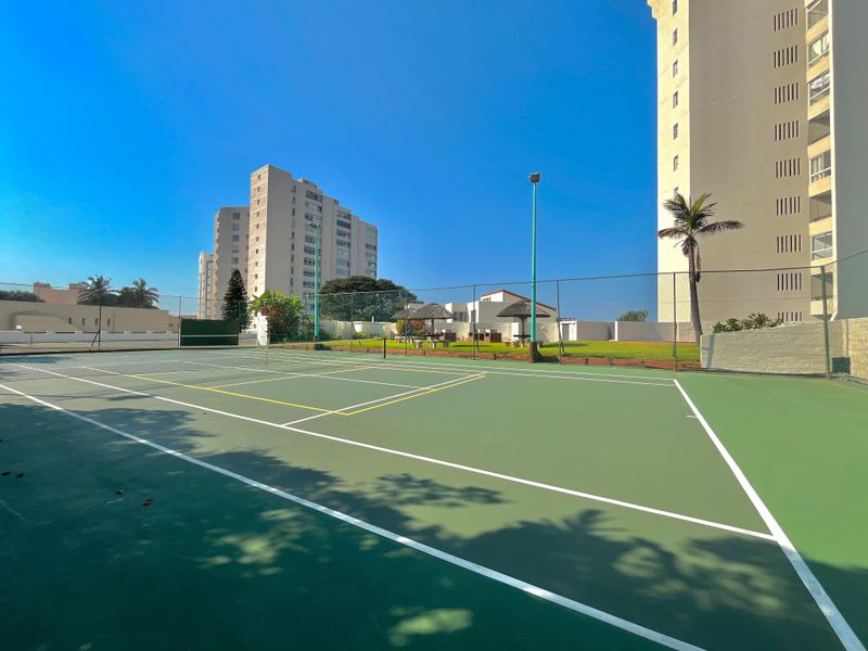 A well-maintained outdoor tennis court featuring a hard surface, perimeter chain-link fencing, and integrated floodlighting within the apartment complex grounds.