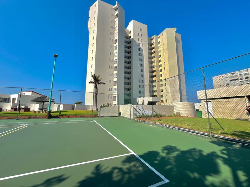 A well-maintained outdoor tennis court situated within the apartment complex grounds, featuring perimeter fencing and adjacent landscaped lawn areas.
