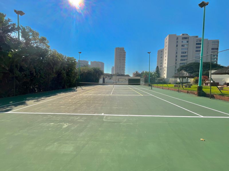Outdoor tennis court facility with perimeter fencing and floodlights, situated within an apartment complex.