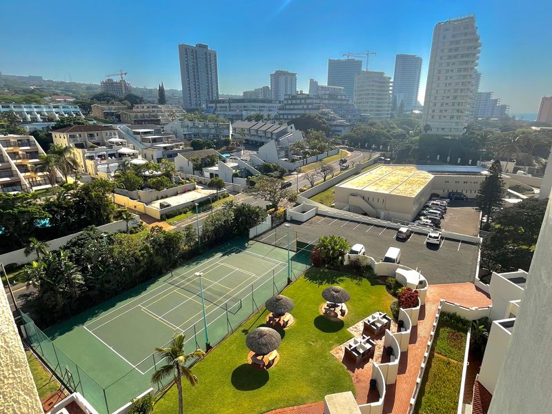 Elevated view overlooking the apartment complex grounds, featuring a swimming pool, tennis court, landscaped gardens, and surrounding urban cityscape.
