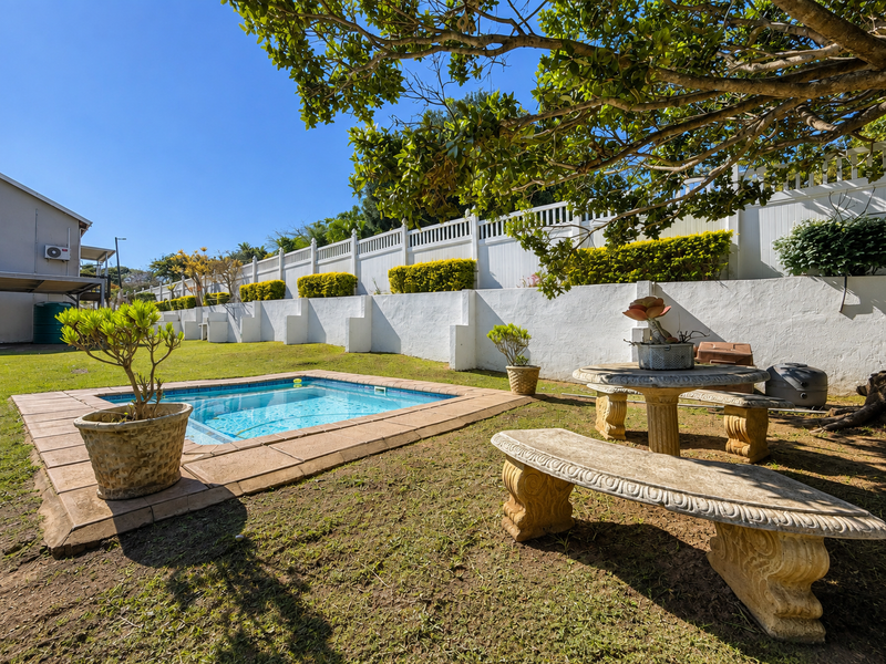 A private backyard featuring a swimming pool with tiled surround, set against a white retaining wall and perimeter fencing in a landscaped garden.
