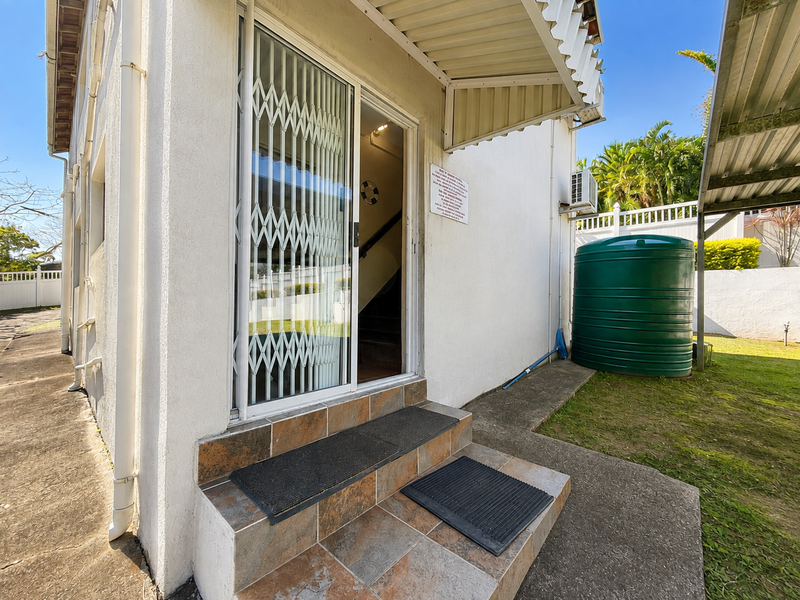Rear exterior view featuring a sliding glass door with security gate, tiled entrance steps, a water storage tank, and an external air conditioning unit.