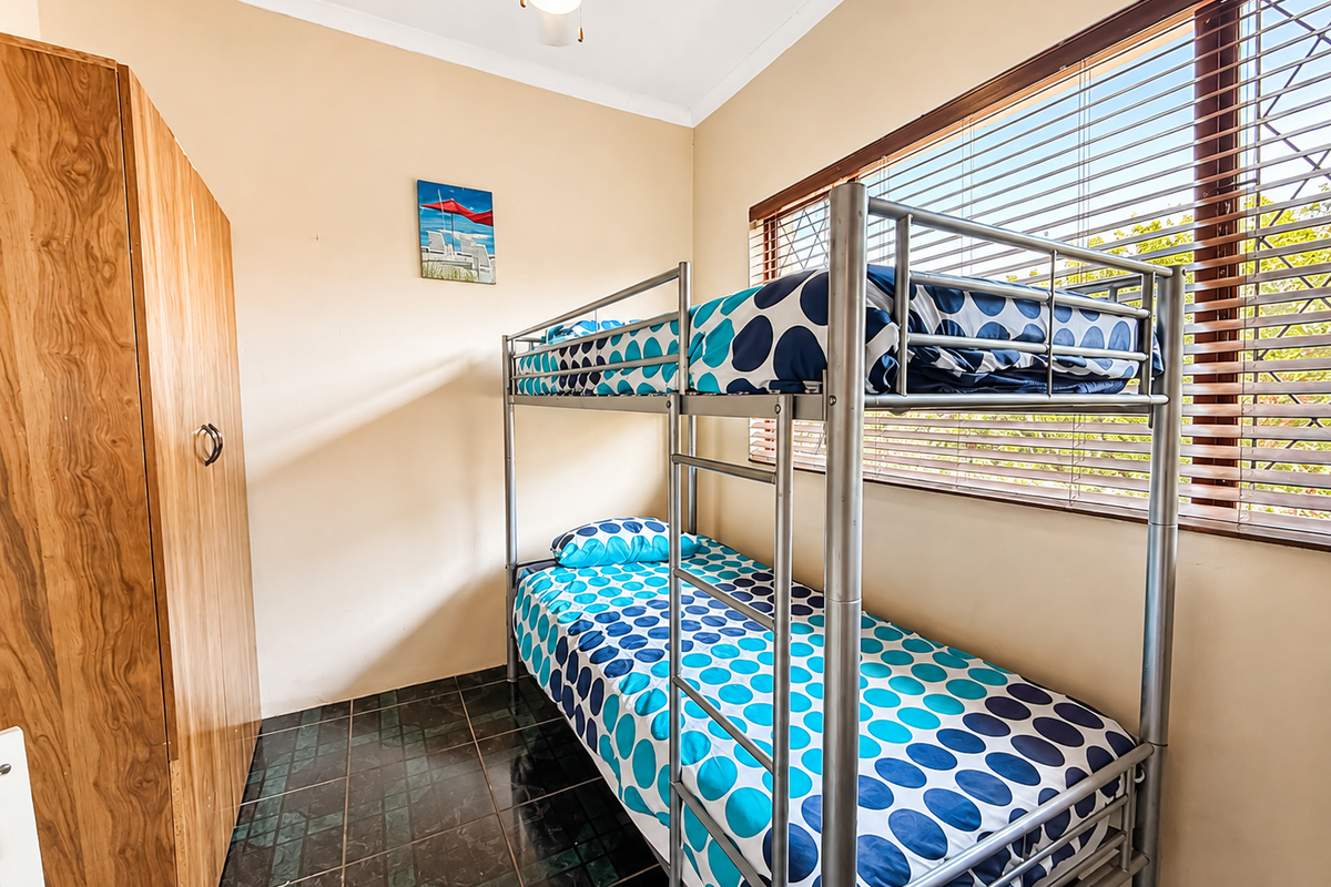 A compact bedroom featuring tiled flooring, a built-in wardrobe, a ceiling fan, and large windows fitted with venetian blinds and security bars.