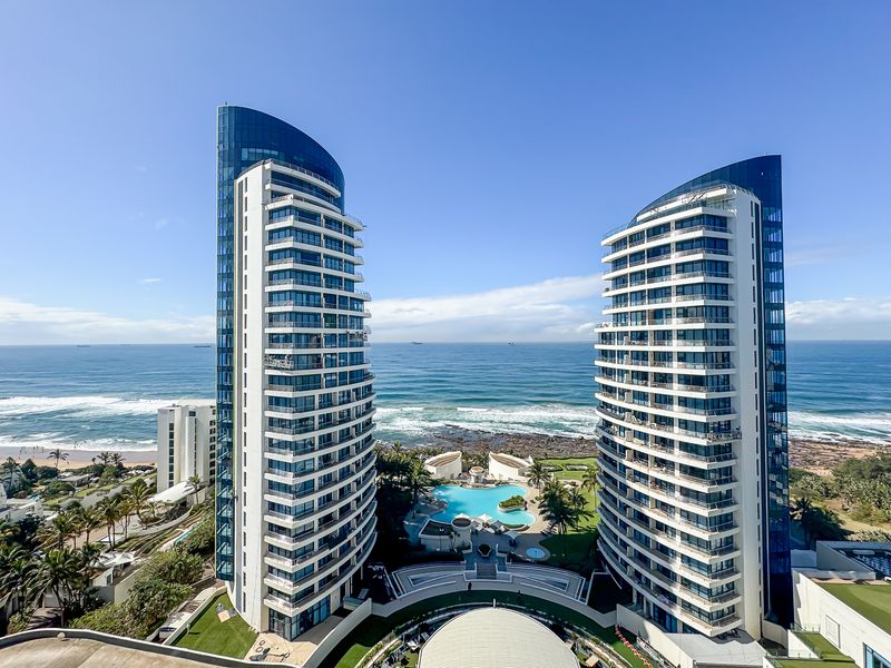Aerial view of a modern luxury apartment complex featuring twin high-rise towers, a central swimming pool, and direct oceanfront access.