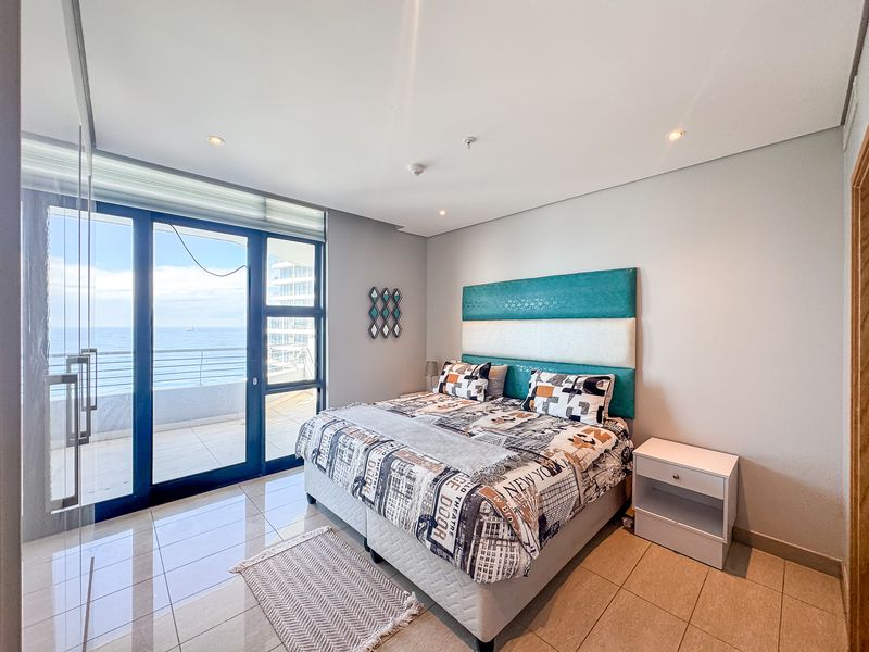 A bright bedroom featuring tiled flooring, recessed lighting, and a large sliding glass door providing direct access to a balcony with ocean views.