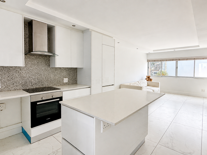 Modern open-plan kitchen and living area featuring sleek white cabinetry, a stone island, integrated appliances, and bright tiled flooring throughout.