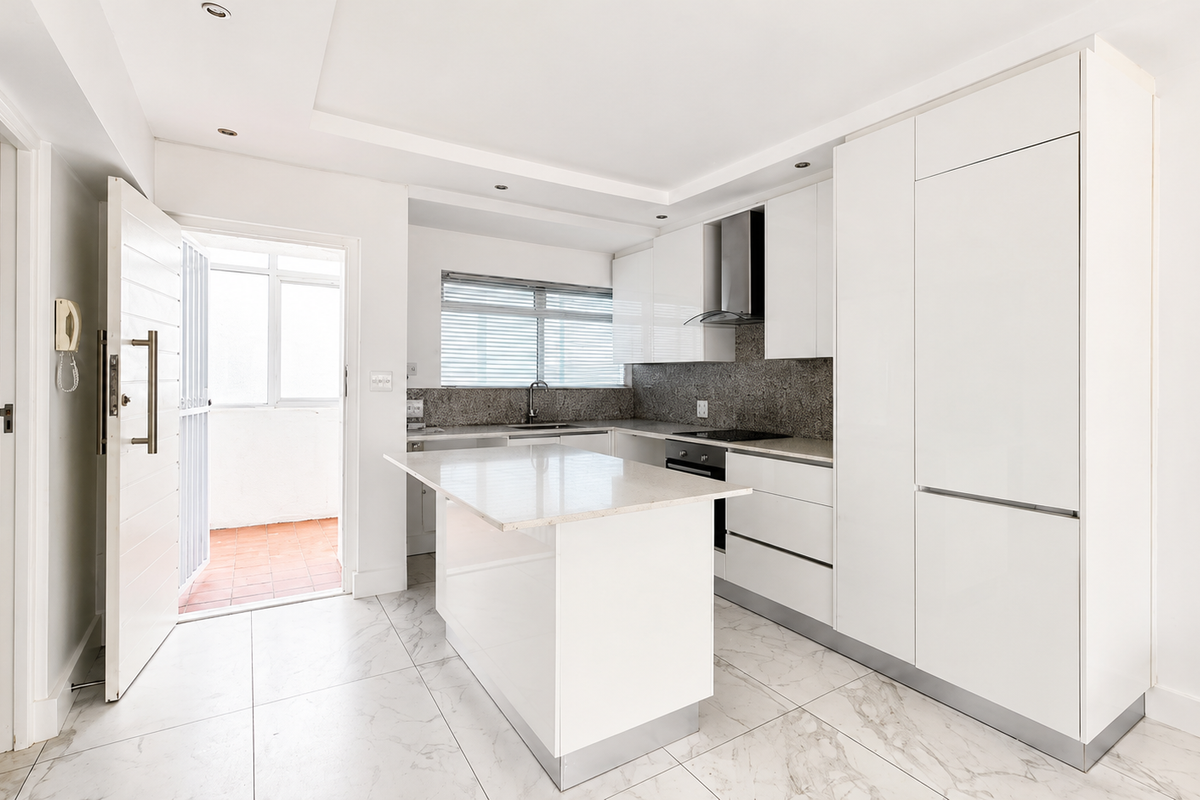 Modern, minimalist kitchen featuring white high-gloss cabinetry, stone countertops, a central island, and integrated appliances, leading to an outdoor balcony area.