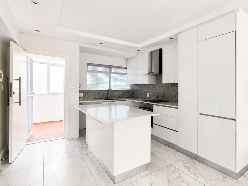 Modern, minimalist kitchen featuring white high-gloss cabinetry, stone countertops, a central island, and integrated appliances, leading to an outdoor balcony area.