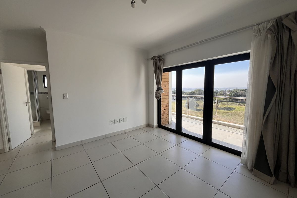 A bright, empty bedroom featuring large sliding glass doors that open onto a balcony with scenic views and neutral tiled flooring throughout.