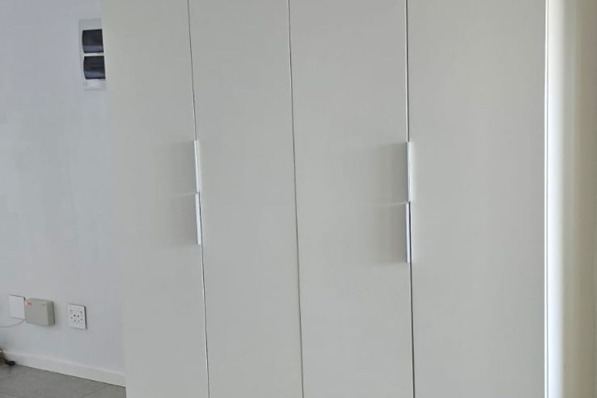 A clean, minimalist bedroom featuring modern white built-in cupboards with sleek handles and neutral tiled flooring.