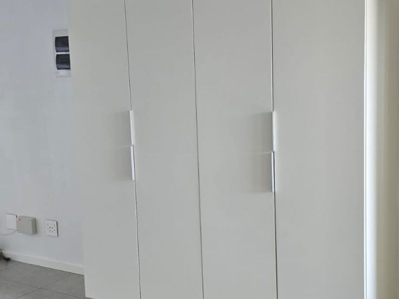 A clean, minimalist bedroom featuring modern white built-in cupboards with sleek handles and neutral tiled flooring.