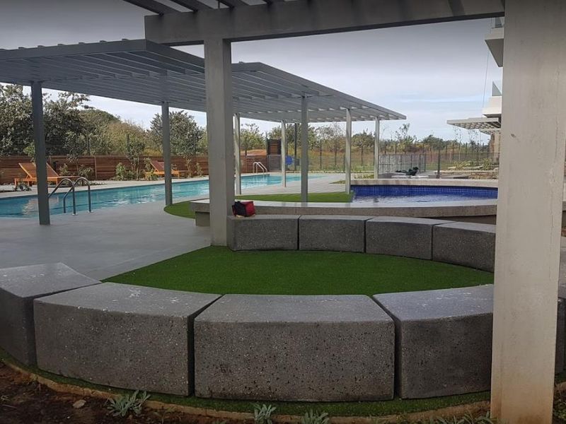 Communal pool area featuring a modern pergola, built-in concrete circular seating, and paved surrounds with artificial turf landscaping.