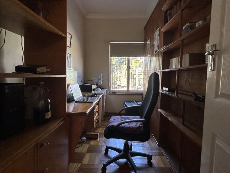 A compact home office featuring built-in wooden shelving, parquet flooring, and a window with security bars, providing a functional workspace.