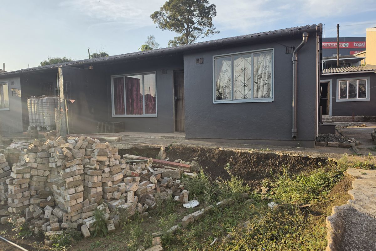Rear view of a single-story house featuring dark painted exterior walls, tiled roofing, and security bars on windows, with significant landscaping and site work required.