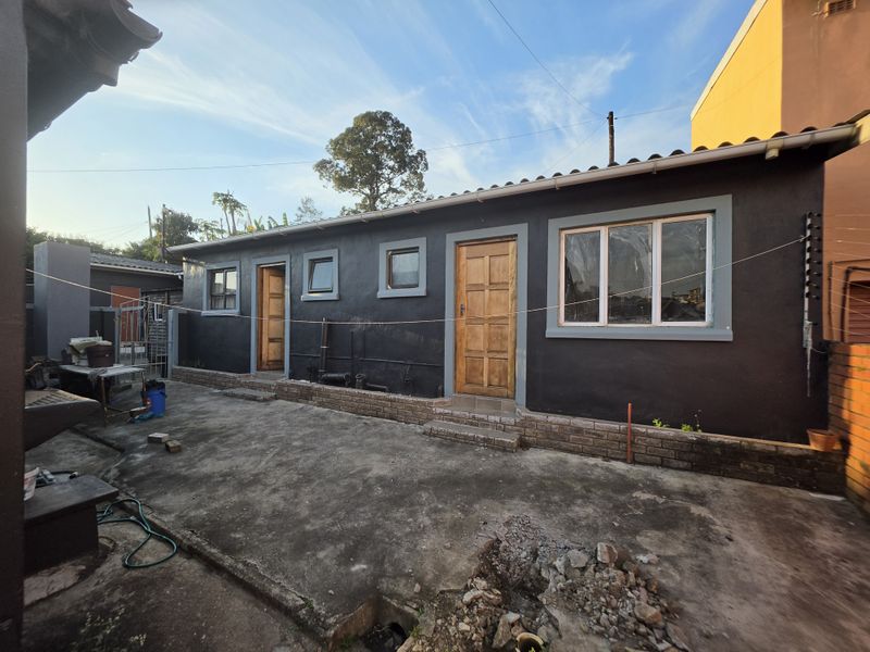 Rear exterior view of the property featuring a concrete patio area, dark painted walls, timber door, and aluminium window frames.