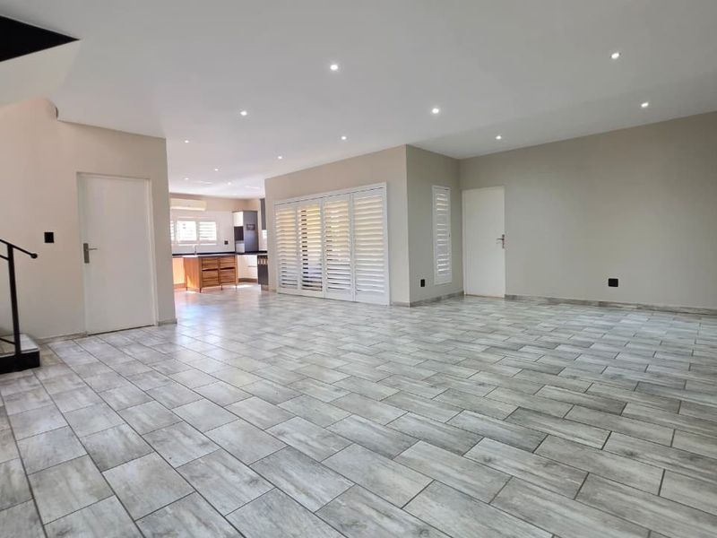 Spacious open-plan living area featuring modern tiled flooring, recessed lighting, and plantation shutters, with an adjoining kitchen and staircase access.