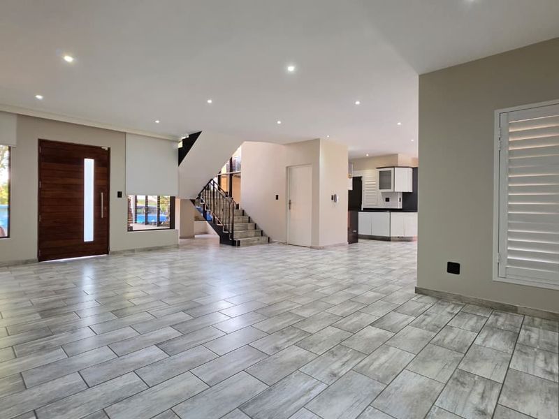 Spacious open-plan living area featuring modern tiled flooring, recessed lighting, a feature staircase, and direct access to the kitchen and entrance.