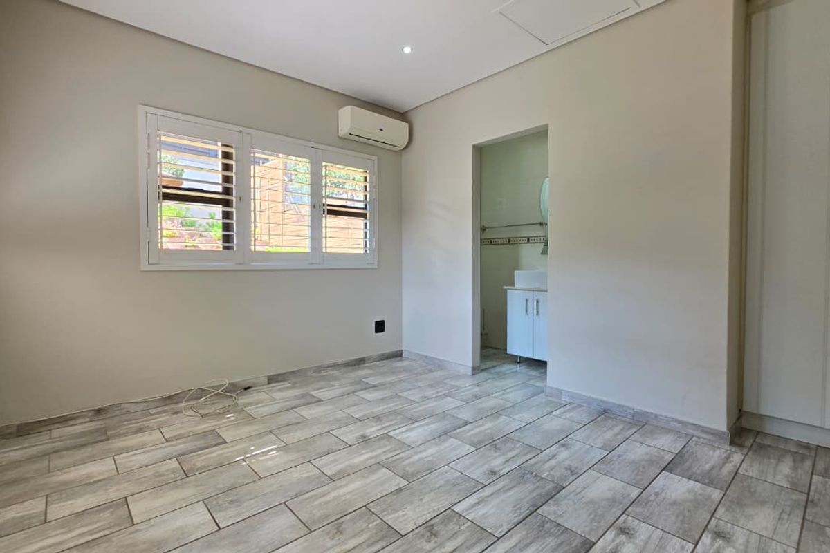 A bright, empty bedroom featuring modern wood-look tiled flooring, built-in cupboards, air conditioning, and shutter blinds, with an adjoining en-suite bathroom.