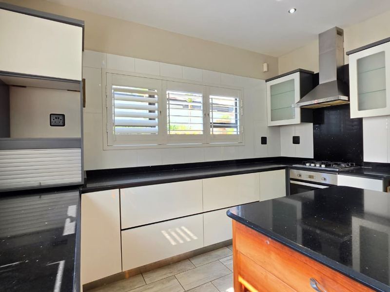 Modern kitchen featuring sleek white cabinetry, black granite countertops, built-in oven and gas hob, stainless steel extractor, and plantation-style shutter blinds.
