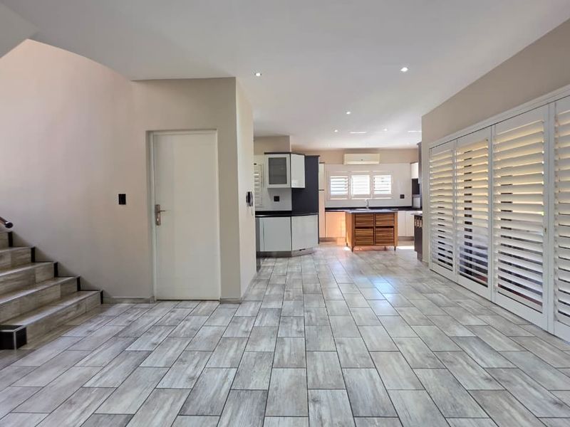 Spacious open-plan living area featuring modern tiled flooring, a staircase with metal railing, and a well-appointed kitchen with built-in cabinetry and plantation shutters.