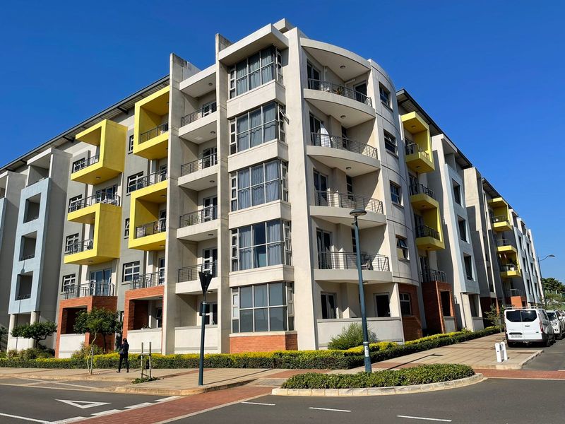 A modern multi-story apartment building featuring a contemporary facade with yellow accents, private balconies, and a paved street-level entrance.