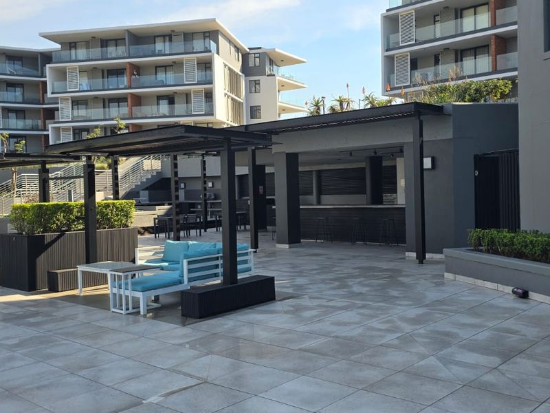 Modern outdoor clubhouse area featuring a tiled patio, pergola structure, and a built-in bar counter, set within a contemporary apartment complex.