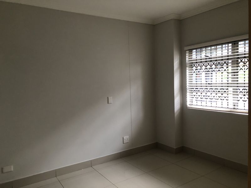 A clean, empty bedroom featuring neutral tiled flooring, fitted venetian blinds, and a security gate on the window.