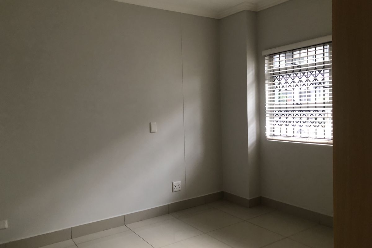 A clean, empty bedroom featuring neutral tiled flooring, a window with security bars and venetian blinds, and well-maintained wall finishes.