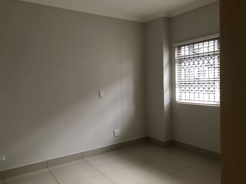 A clean, empty bedroom featuring neutral tiled flooring, a window with security bars and venetian blinds, and well-maintained wall finishes.