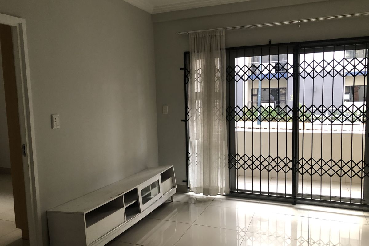 A bright, empty living room featuring neutral tiled flooring, cove cornicing, and a sliding glass door with a security gate leading to a balcony.