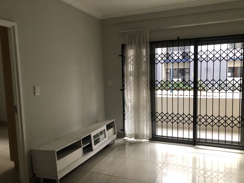 A bright, empty living room featuring neutral tiled flooring, cove cornicing, and a sliding glass door with a security gate leading to a balcony.
