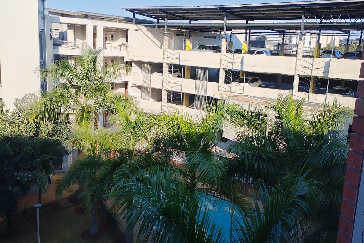 An elevated view of the apartment complex grounds, featuring a swimming pool, landscaped garden area, and a multi-level parking structure.