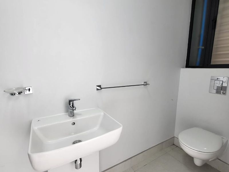 A clean, modern bathroom featuring a white pedestal washbasin, wall-mounted toilet with chrome flush plate, and neutral floor tiling.