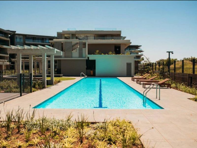 A well-maintained communal swimming pool area featuring a lap pool, paved surround, and a modern pergola structure within an apartment complex.