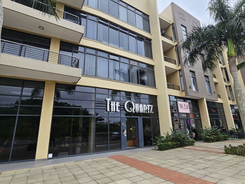 Front elevation of The Quartz apartment complex featuring a modern glass facade, ground floor commercial units, and paved pedestrian access.