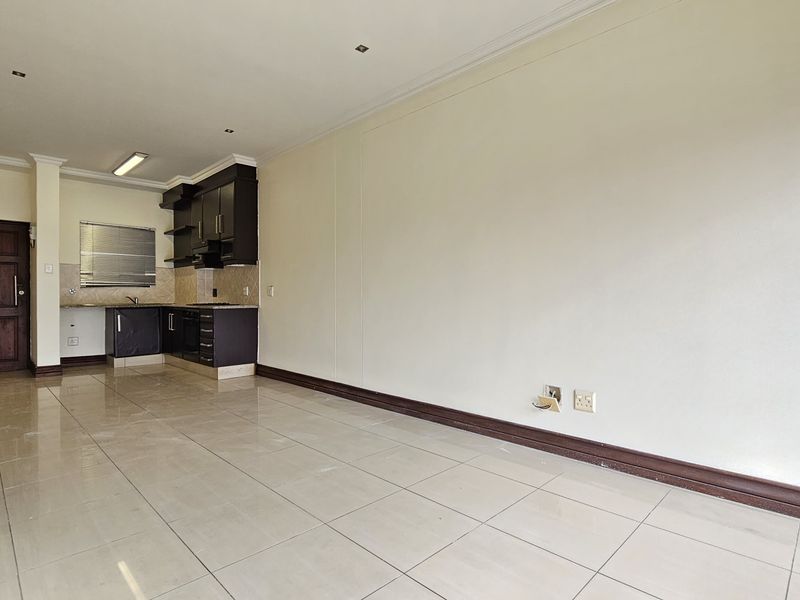 An open-plan living and kitchen area featuring tiled flooring, dark built-in cabinetry, and integrated kitchen appliances in a vacant apartment setting.
