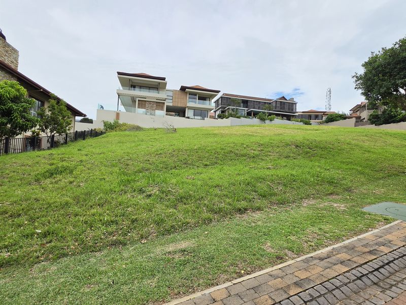 A sloped residential vacant land plot featuring established grass, a paved driveway edge, and surrounding perimeter fencing in a developed estate setting.