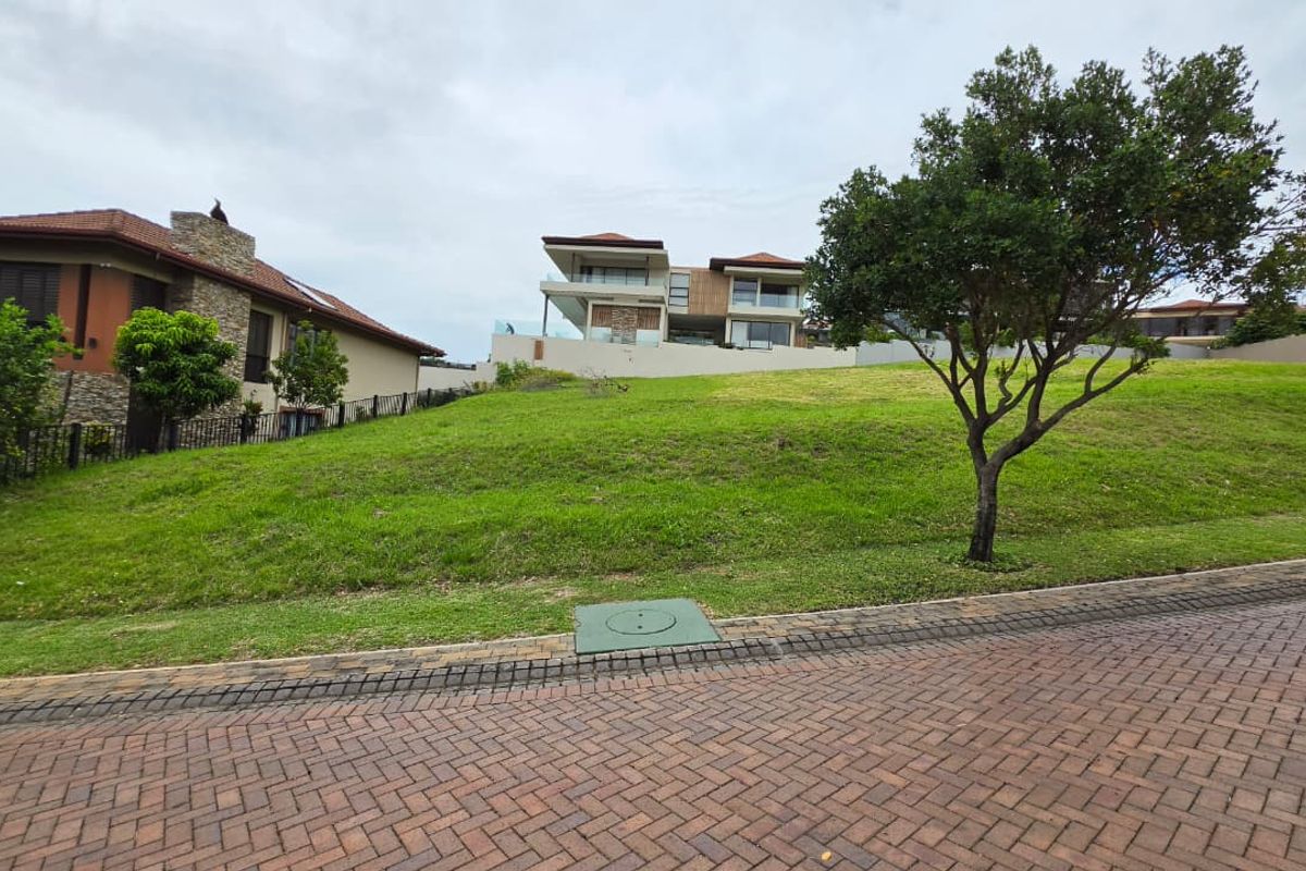 A vacant residential plot featuring a gentle upward slope, manicured lawn, and paved street access in a developed estate setting.