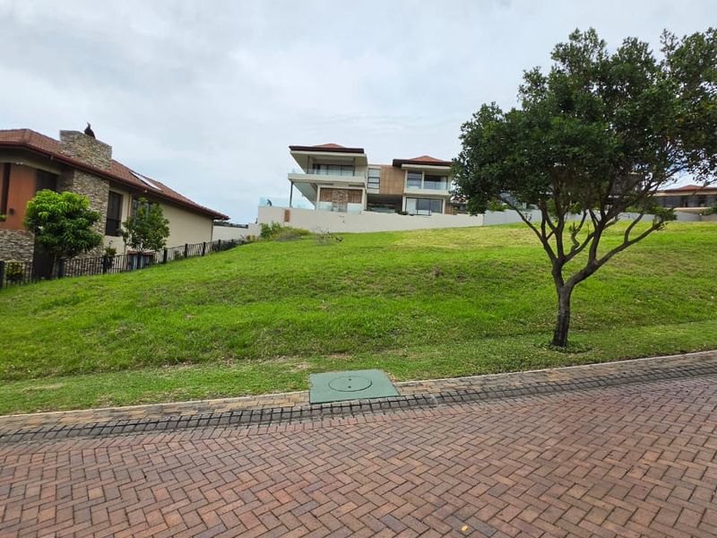 A vacant residential plot featuring a gentle upward slope, manicured lawn, and paved street access in a developed estate setting.