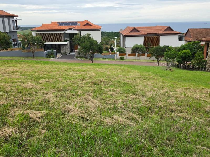 A vacant residential plot featuring a gentle slope, situated in a developed estate with established neighboring homes and street access.