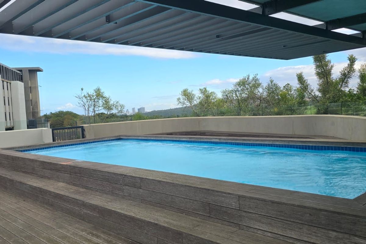 A modern raised swimming pool area featuring timber decking, a louvred pergola, and a glass balustrade with scenic views.