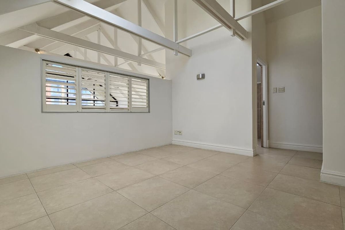 An empty bedroom featuring high ceilings with exposed white roof trusses, tiled flooring, and plantation shutters, offering a bright and clean living space.