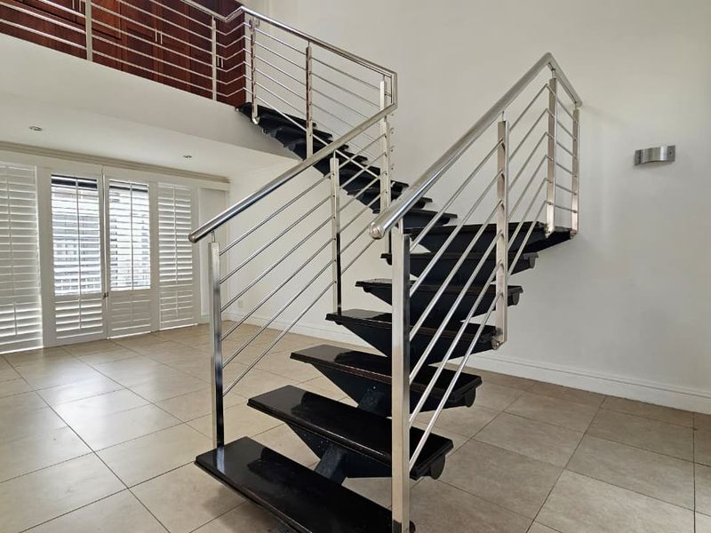 Spacious open-plan living area featuring a modern staircase with stainless steel balustrades, tiled flooring, and plantation shutters, offering a bright and airy atmosphere.