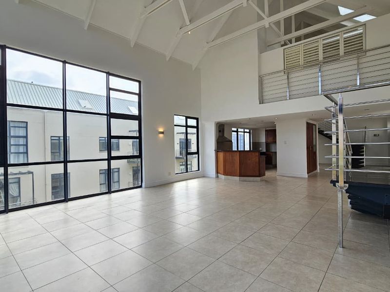Spacious open-plan living area featuring vaulted ceilings, exposed trusses, large windows, and a mezzanine level with a modern stainless steel balustrade.