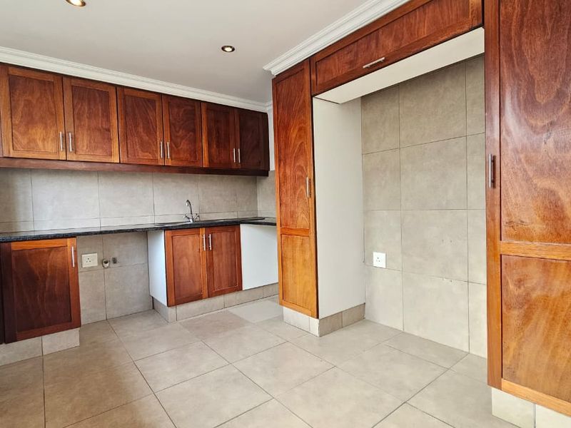 A clean, empty kitchen featuring warm wooden cabinetry, granite countertops, a stainless steel sink, and neutral tiled finishes throughout.
